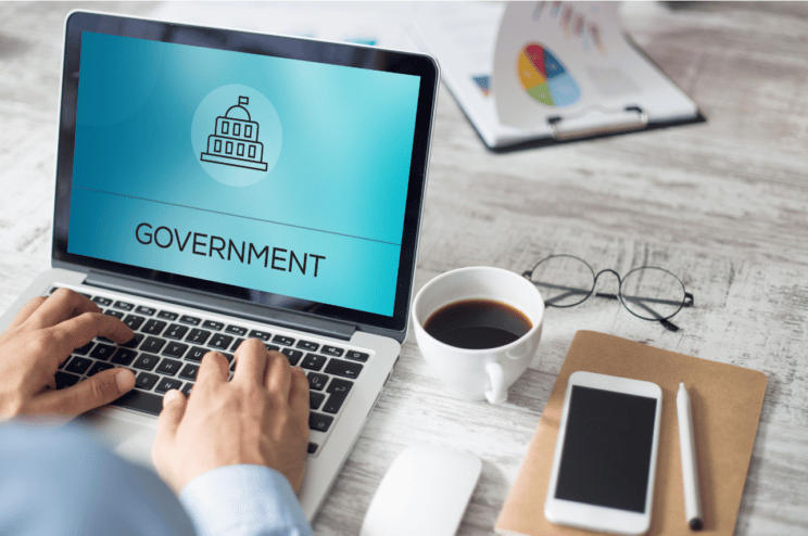 A person types at a laptop, with a coffee and smartphone alongside them. The laptop screen displays a graphic which reads "government".