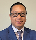 Professional headshot of a man in a blue suit and tie, wearing glasses, against a neutral background.