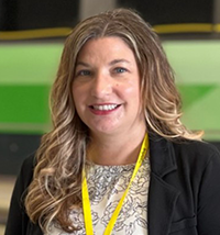 Smiling woman with wavy blonde-brown hair, wearing a black blazer and patterned blouse, with a yellow lanyard in a professional setting.